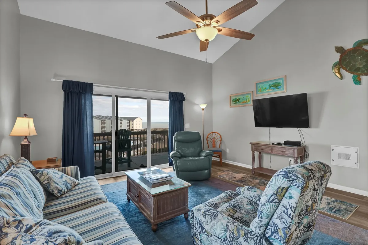 A cozy, open living room with a plush sofa and armchair facing a smart TV, accented by an inviting sliding glass door that reveals a private balcony and glimpses of the ocean beyond. The sunset light pours in, creating an atmosphere ideal for relaxation and socializing.