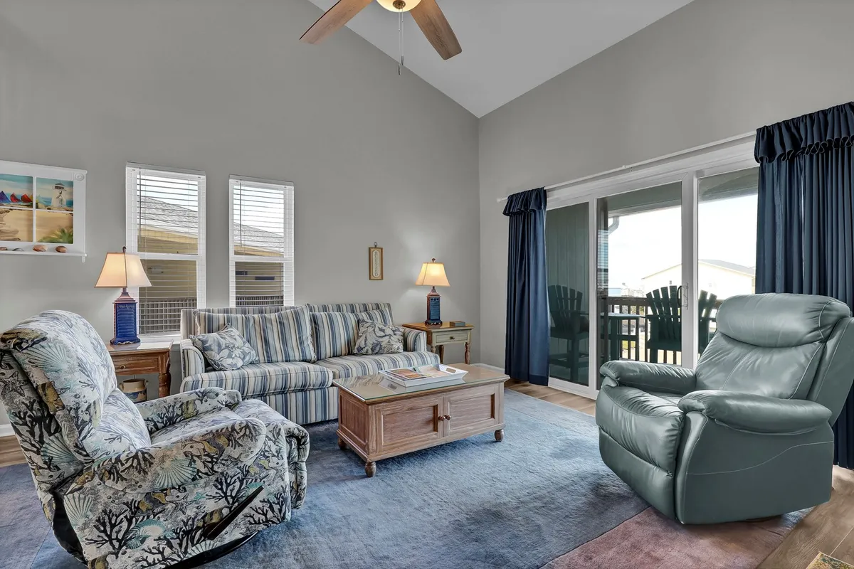 A sunlit living room with a striped sofa, overstuffed armchair, coffee table, and large windows offering an outdoor view. The space is open, cozy, and inviting, with hard flooring and minimal clutter, making it practical for guests traveling with pets.