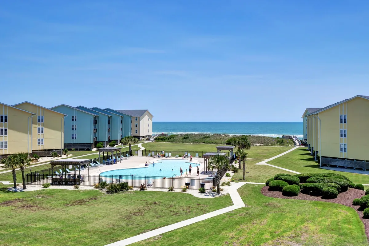 A scenic exterior shot of the swimming pool complex with ocean views, green lawns for lounging or picnicking, and nearby buildings. The space feels open and welcoming, ideal for group gatherings or unwinding after a day exploring Surf City.
