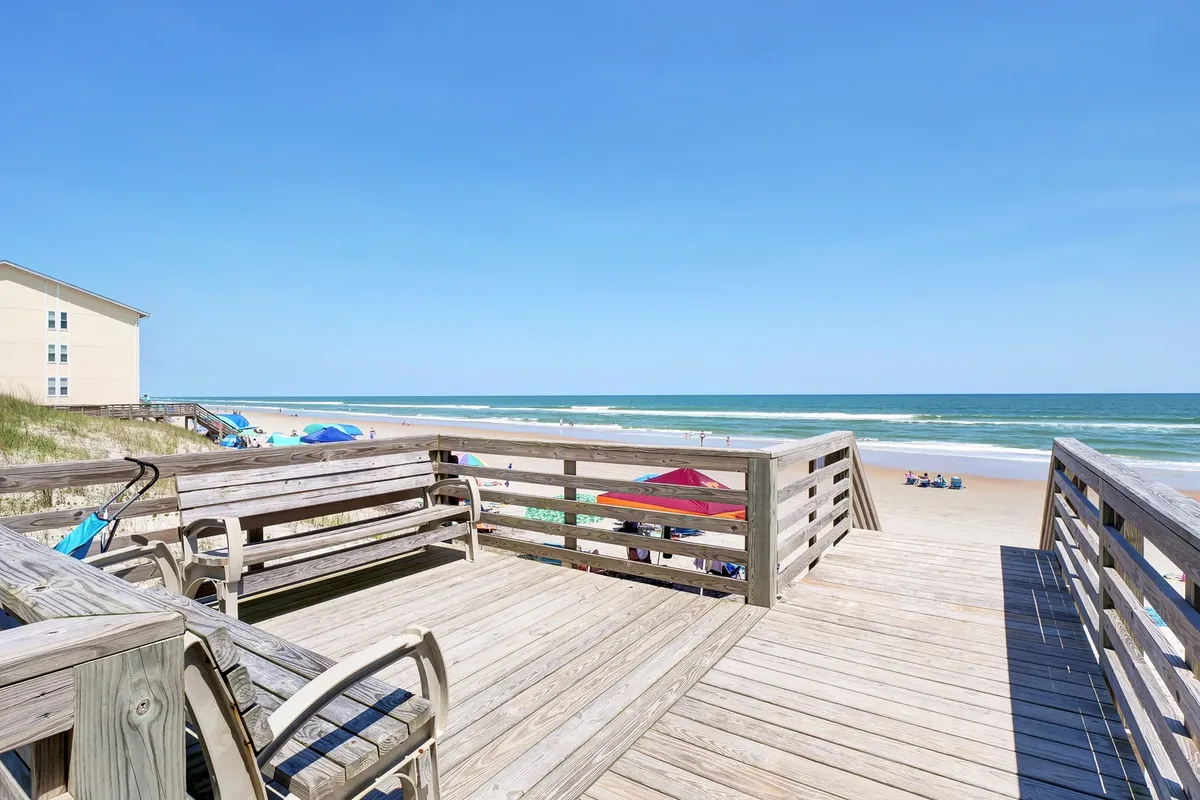 A wooden deck overlooking the ocean, dotted with beach umbrellas and lounge chairs, set against a serene, expansive shoreline. The scene conveys easy access to the beach for morning walks and sunset strolls, capturing the spirit of daily outdoor adventures with pets.