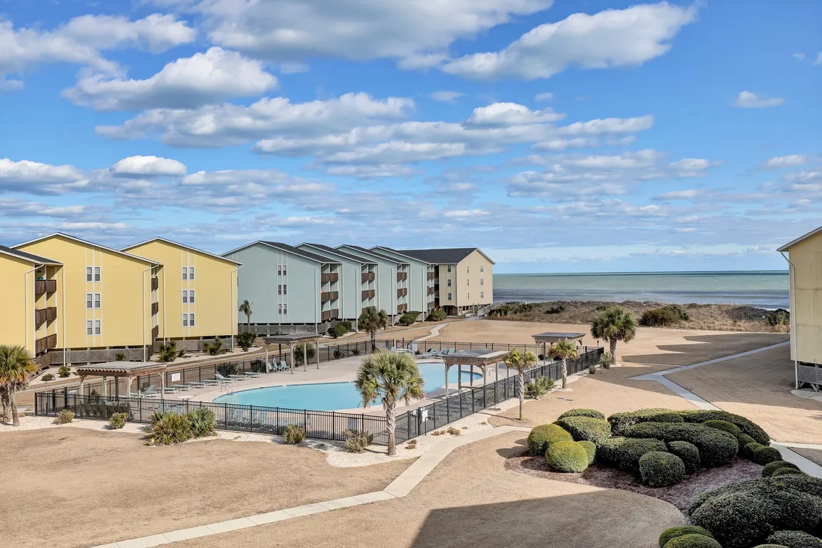 The hero image showcases a sparkling swimming pool surrounded by lush, landscaped gardens, with vibrant, colorful condo buildings framing the scene. Beyond the pool, there are sweeping views of the Atlantic Ocean, capturing the relaxed coastal atmosphere. Guests can be seen lounging by the pool and enjoying the sunset, evoking a sense of leisure and seaside charm.