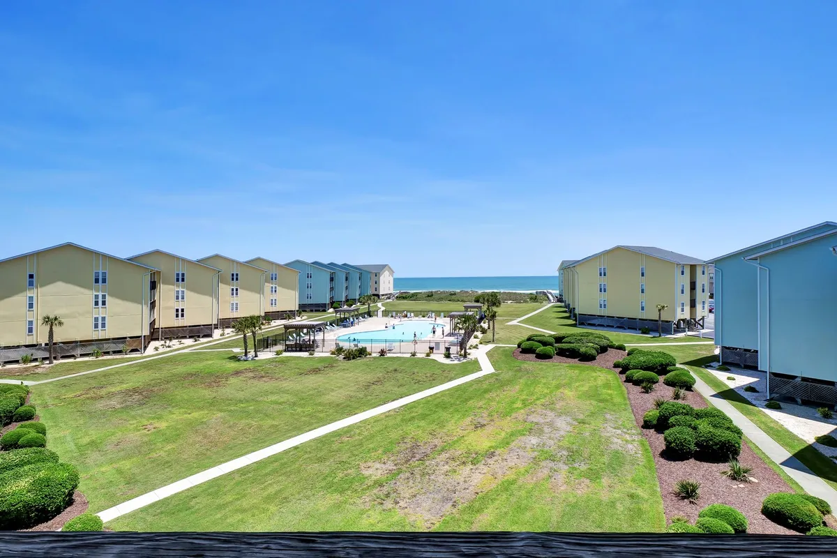A sunlit outdoor community pool sits at the center of the property, bordered by neatly landscaped grounds and residential buildings. Lounge chairs are visible around the pool, inviting guests to relax and enjoy the atmosphere. The surrounding environment is tranquil, with soft natural light accentuating the water's sparkle.