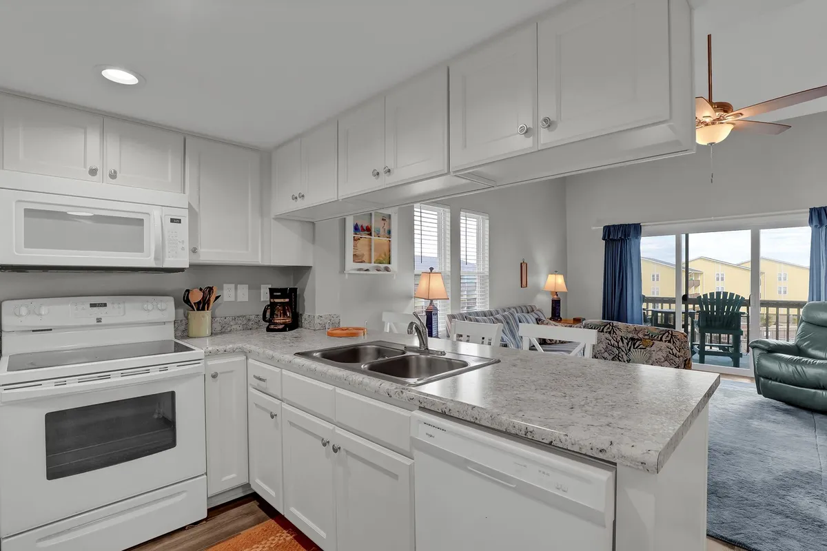A welcoming kitchen scene featuring a spacious island, bright white cabinetry, and a seamless connection to a cozy living area with chairs and lamps. The open layout encourages cooking, entertaining, and easy flow between spaces.