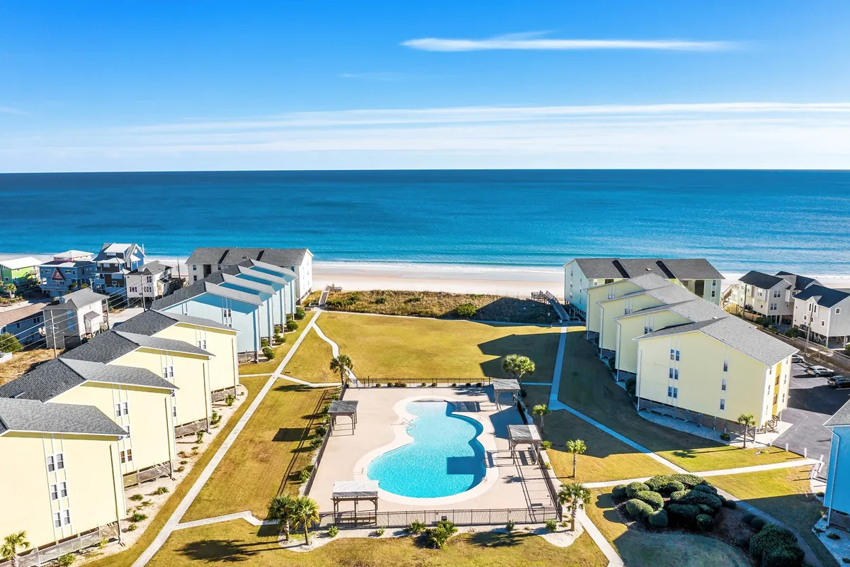 The exterior scene features a swimming pool, colorful townhouses, green lawns, and the beachfront, with space for sunbathing, picnicking, and enjoying the outdoors with neighbors.