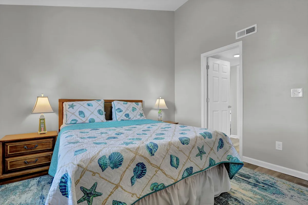 A serene bedroom featuring a bed with a seashell-patterned quilt, a nightstand with a lamp, and coastal decorative elements. The room is uncluttered and bright, suggesting it’s easy to maintain and comfortable for everyone, including pets.
