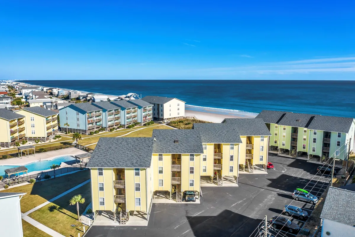 A panoramic exterior view capturing the pool, multi-story condo buildings, and expansive ocean. The scene suggests a lively, resort-like atmosphere where families or friends can gather for poolside barbecues, walks on the beach, or exploring the local area together.