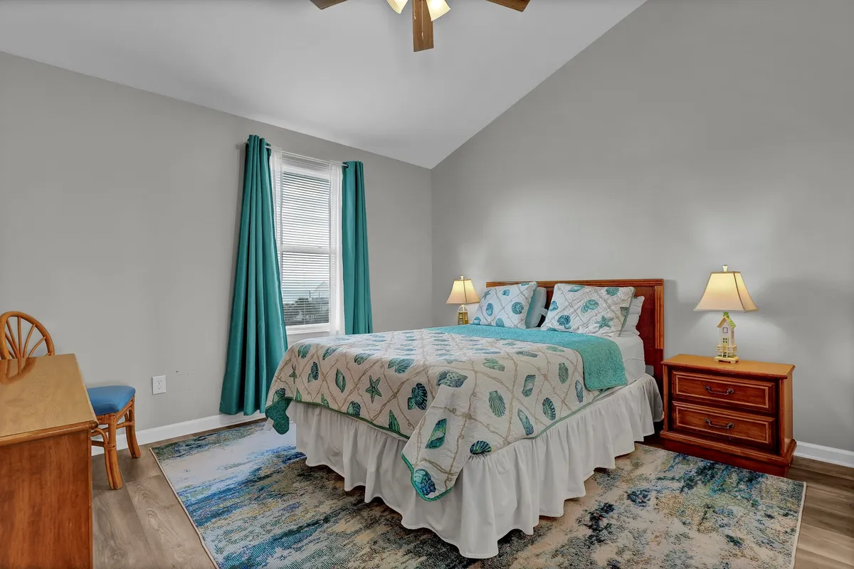 A serene master bedroom with a plush bed adorned with decorative bedding, a nightstand with a soft-glowing lamp, a desk and chair by the window, and curtains that gently filter the daylight. The arrangement creates a cozy and restful environment for unwinding after a busy day at the beach.