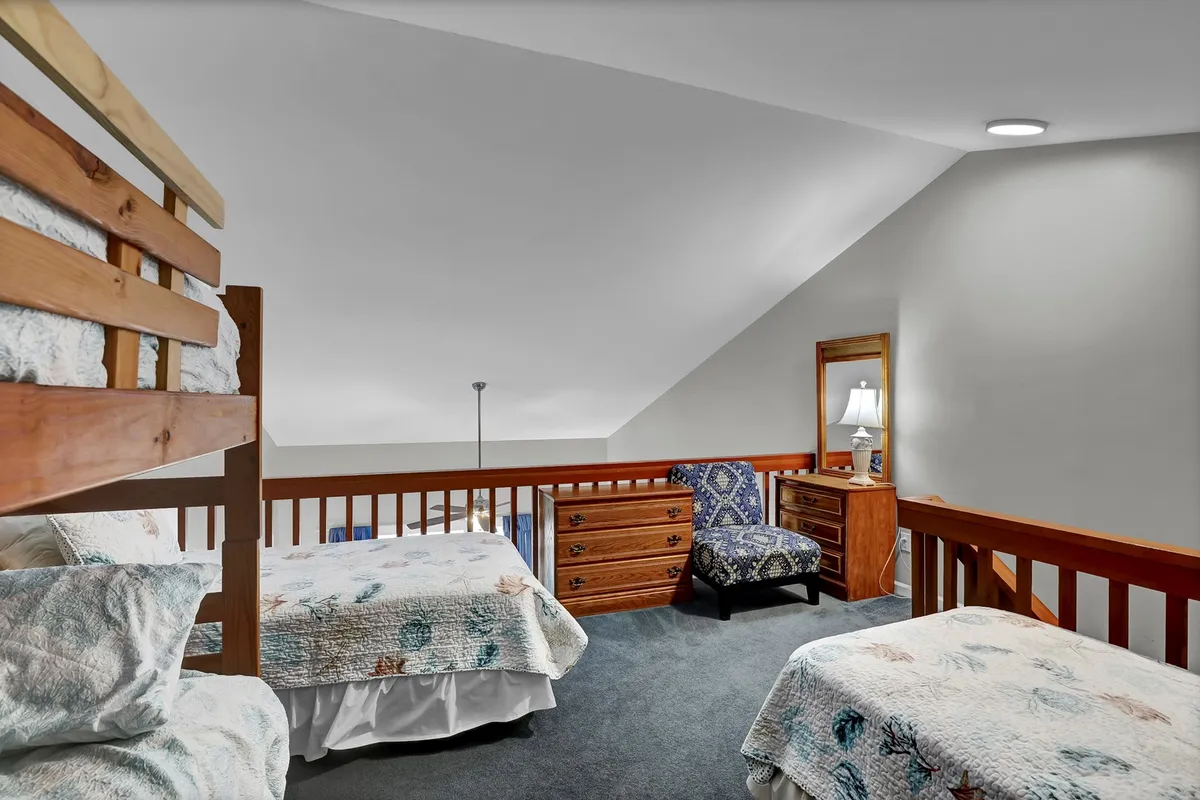A versatile loft or secondary sleeping area featuring a bunk bed and two single (twin) beds, complemented by an armchair and a wooden dresser. The space feels inviting and perfect for group stays, with plenty of room for kids or friends to share stories or play games.