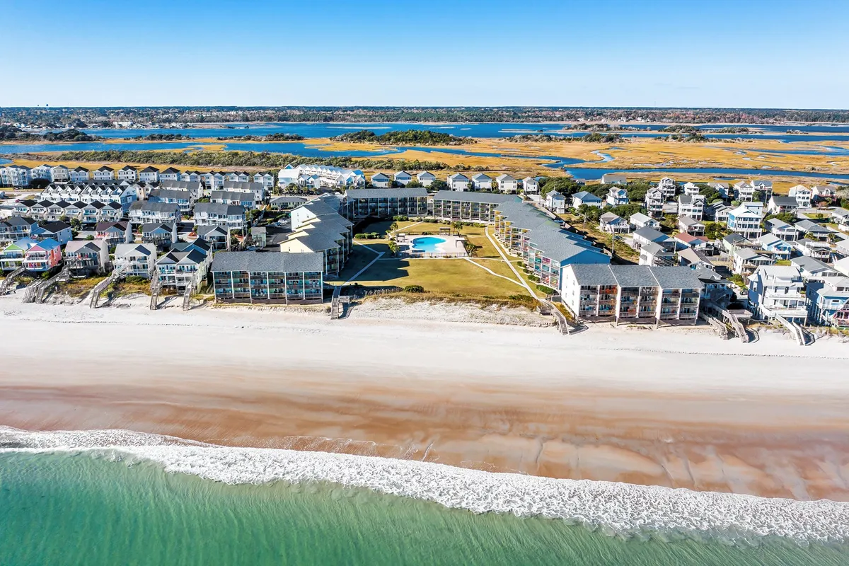 A wide exterior view showing the beachfront, the sparkling community swimming pool, and nearby surrounding houses. The scene features families and groups enjoying the sandy shore, sunbathing by the pool, and socializing in a relaxed, beachside environment.