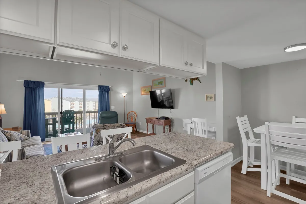 A bright, open-plan kitchen and dining area with a kitchen sink, white dining table, comfortable living area with a couch, and a large window letting in ample natural light. The surfaces appear durable and easy to clean, making the space welcoming and practical for families with pets.