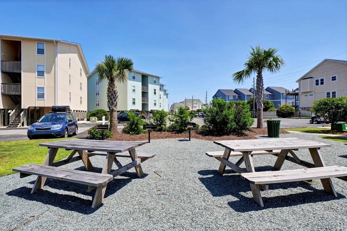 A sunny outdoor communal area featuring picnic tables shaded by palm trees, surrounded by low-rise condo buildings. The space is vibrant and inviting, ideal for outdoor gatherings, walks, or playtime with pets in a relaxed, social setting.